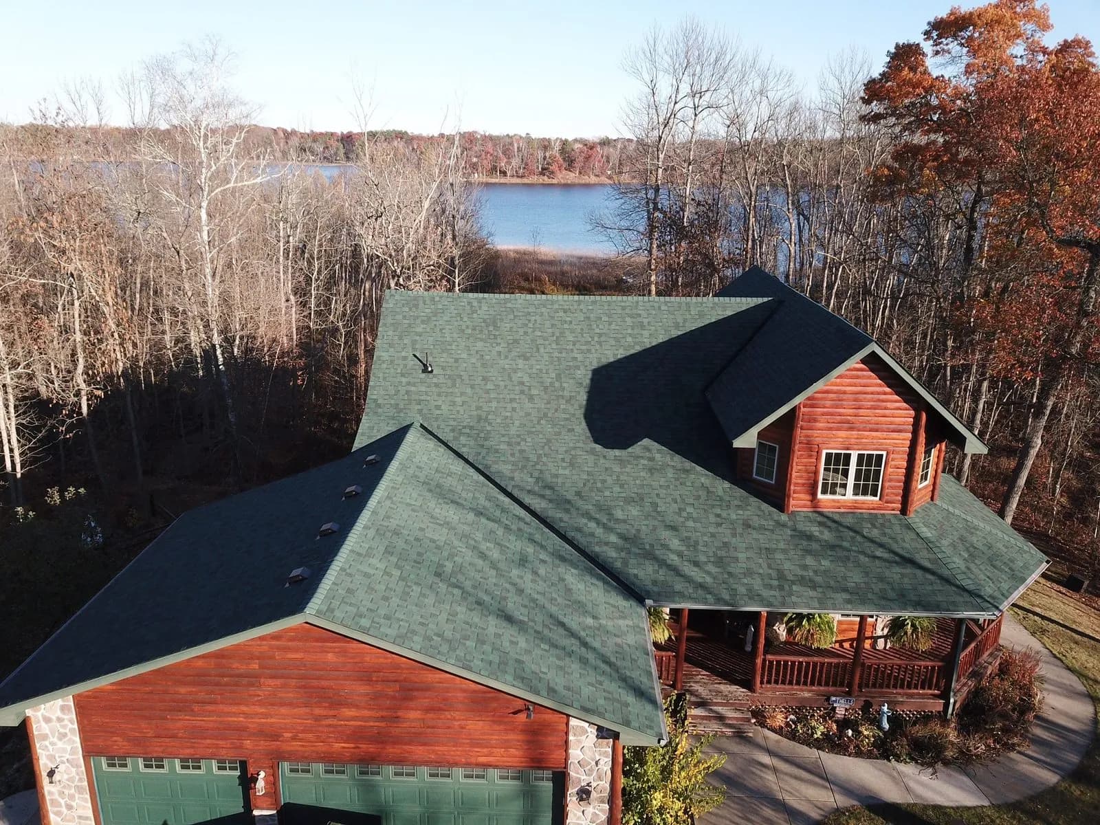 Aerial view of a lakeside log home with a new teal green shingle roof surrounded by autumn trees