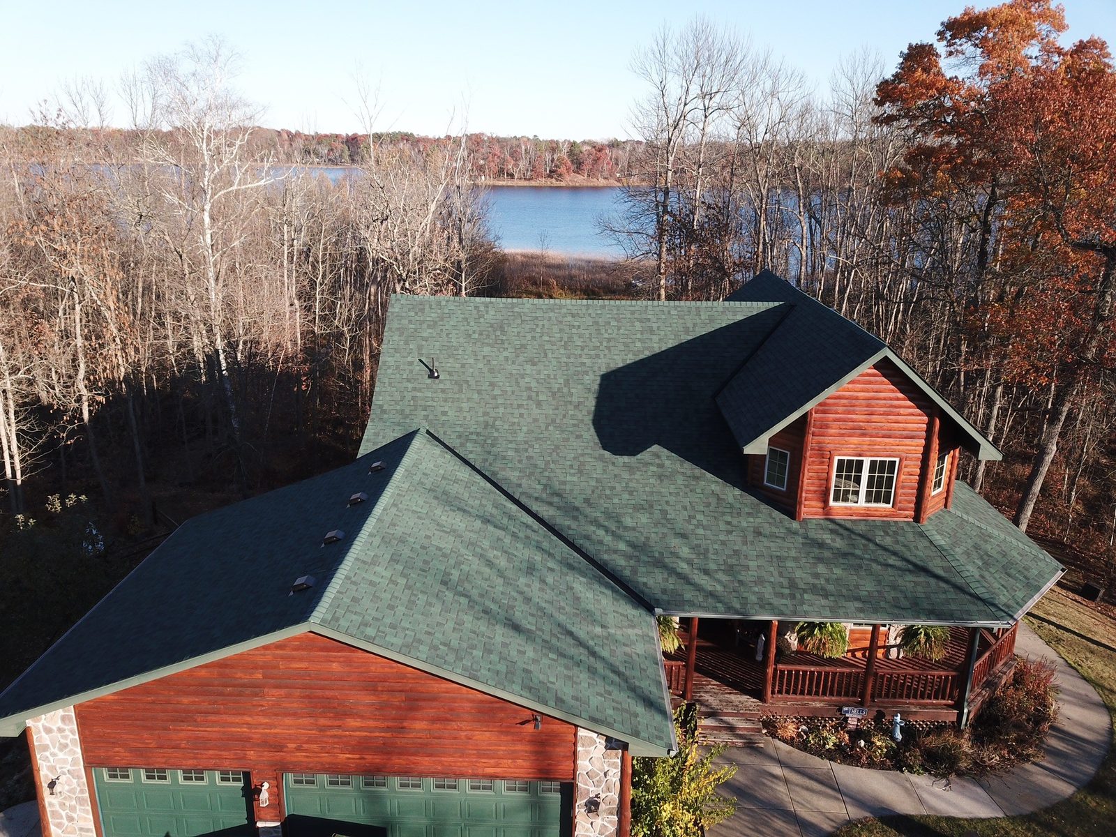 Aerial drone view of a lakeside log home with a new teal green shingle roof surrounded by autumn trees