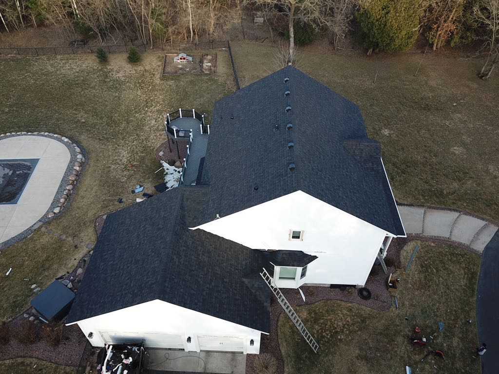 Aerial drone view of a large home with a dark shingle roof and a swimming pool
